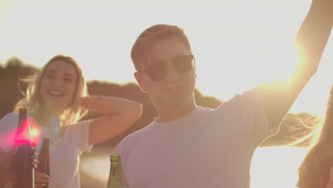 Young-boy-in-white-T-shirt-and-black-fashionable-sun-glasses-is-moving-his-hands-and-dancing-with-his-best-friends-on-the-open-air-party-with-beer.-He-enjoys-this-summer-evening-at-sunset.